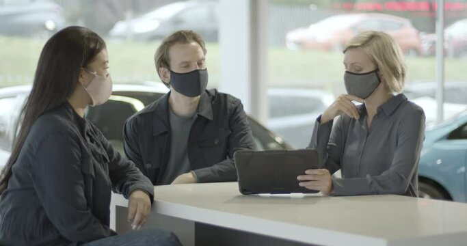 Saleswoman advising couple on car purchase in vehicle dealership showroom wearing face mask