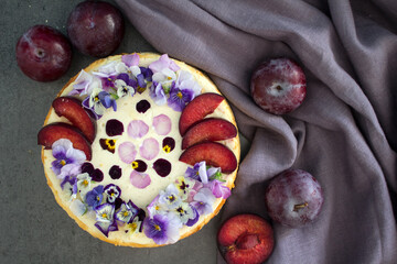 Cheesecake decorated with red plums and edible flowers. Fabric texture background. Top view photo of homemade dessert. 
