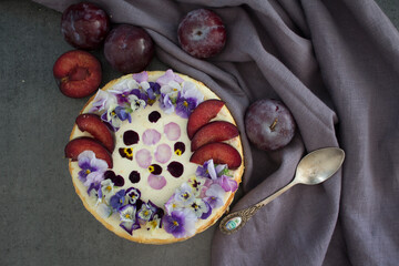 Cheesecake decorated with red plums and edible flowers. Dark background. Top view photo of homemade dessert. 
