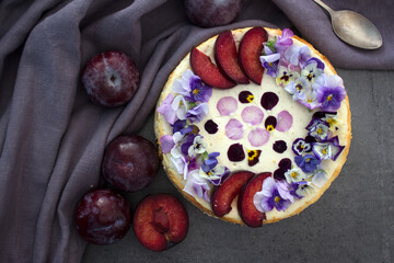 Cheesecake decorated with red plums and edible flowers. Dark background. Top view photo of homemade dessert. 
