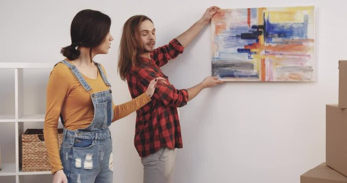 Young beautiful girl instructs her husband where best to attach picture in new apartment. Moving. Arrangement of new house.