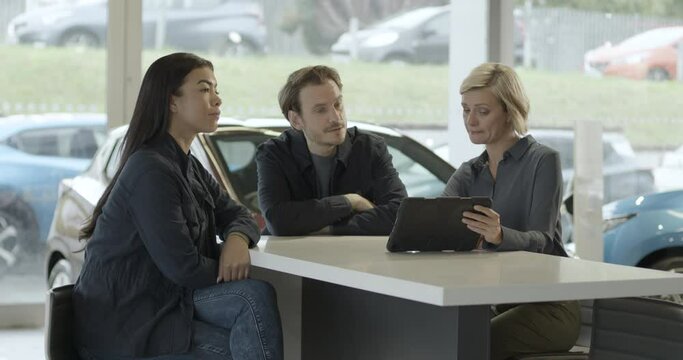 Saleswoman advising couple on car purchase in vehicle dealership showroom using digital tablet