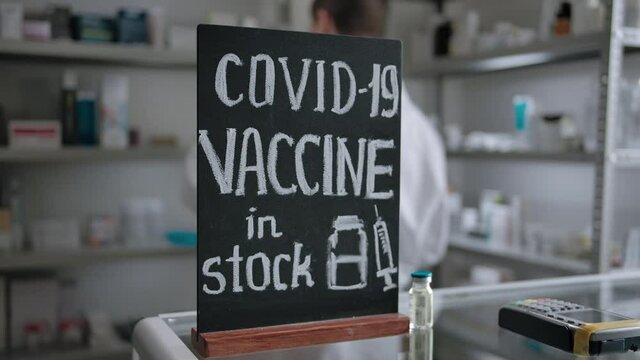 A chalk plaque with the inscription about the presence of a vaccine for coronavirus covid-19 with unrecognizable pharmacist at the background in the interior of the drug store.