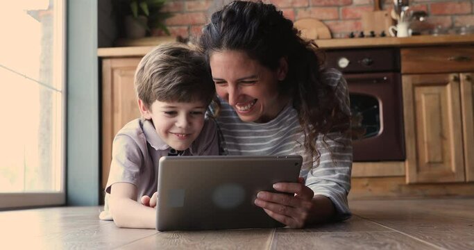 Mom and little son spend free funny time together lying on warm wooden floor at modern house using tablet laughing while watching videos having fun on internet. Modern tech, parental control concept