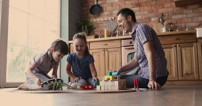 Dad with little 6s 7s children pretty son and daughter rest sit on warm floor in modern cozy kitchen enjoy game together playing wooden bricks and railroad. Funny weekend, hobby, toy store ad concept