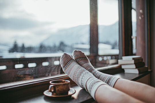Feet in woollen socks by the Alps mountains view. Woman relaxes by mountain view with a cup of hot drink. Close up on feet. Winter and Christmas holidays concept.