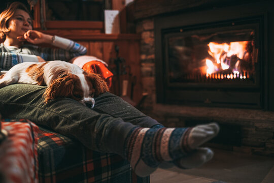 Unrecognizable Woman Feet In Woollen Socks Relax By Cozy Burning Fireplace With Cute Puppy Sleeping By. Snug evening by the warm fire during Christmas season in Switzerland Alps Mountain Chalet