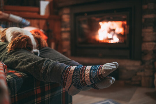 Unrecognizable Woman Feet In Woollen Socks Relax By Cozy Burning Fireplace With Cute Puppy Sleeping By. Snug evening by the warm fire during Christmas season in Switzerland Alps Mountain Chalet