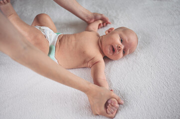 Mom doing exercises for baby in diaper. Cute emotional funny newborn infant little boy in crib.