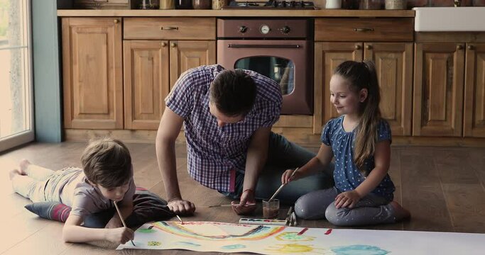 Dad with little son and daughter sit on floor in cozy kitchen spend free leisure time paint with paints on paper, create rainbow colorful picture. Hobby and fun with kids at home, development concept