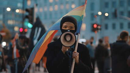 Young woman with face mask speaking into the megaphone while holding rainbow flag in crowd. High...