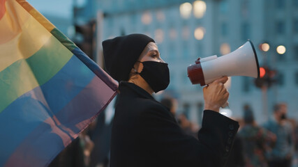 Young woman with face mask speaking into the megaphone while holding rainbow flag in crowd. High...
