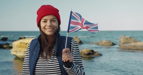 Beautiful girl tourist, exchange student holding in her hands flag of Great Britain and smiles looking at camera. - Powered by Adobe