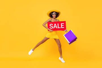 Fototapeta na wymiar Fun portrait of smiling African American woman jumping in mid air while holding red sale signage and shopping bags in isolated studio yellow background