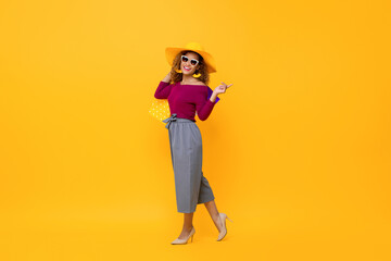Full length portrait of happy African American woman walking carrying shopping bags while pointing on the side in isolated studio yellow background