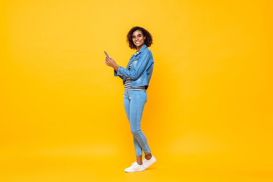 Full Length Portrait Of Young Smiling African American Woman Looking At The Camera While Holding Mobile Phone In Isolated Studio Yellow  Background