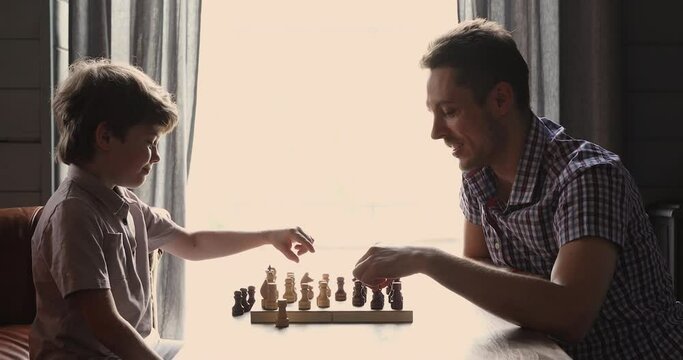 Side view cute little 6s son and young father play chess seated indoor. Concentrated boy develop strategy, planning, foresight skills, engaged in board game with parent at home, entertainment concept