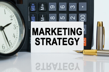 On the table there is a clock, a pen, a calculator and a business card on which the text is written - MARKETING STRATEGY