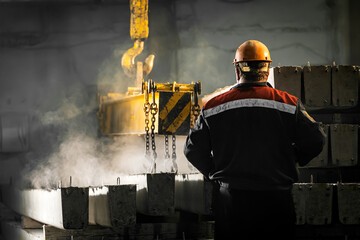 A male worker controls the production process in a factory as a crane moves a reinforced concrete product with holes. Reinforced concrete pillars secured with metal hooks and chains 