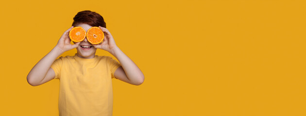 Small caucasian ginger boy is covering his eyes with oranges posing in a t-shirt on a yellow studio wall with free space © Strelciuc