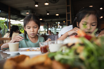 Two asian child girls eating delicious fried fish and salad in the restaurant with family.