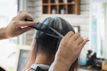 Fototapeta premium Asian young man being cut from long hair to short hair with scissor by professional barber in barbershop.