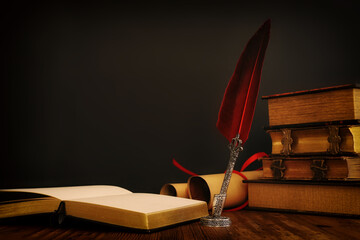 Old feather quill ink pen with inkwell and old books over wooden desk in front of black wall background. Conceptual photo on history, fantasy, education and literature topic.