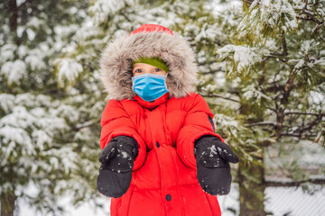 Fototapeta na wymiar Happy boy plays with snow wearing a medical mask during COVID-19 coronavirus. Cute kid throwing snow in a winter park. Happy winter holidays. Winter fashion