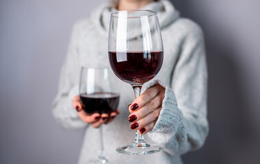 A woman in a white sweater is holding two glasses of red wine and holding out one of them. Close up