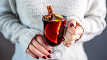 A woman in a white warm sweater is holding a transparent glass cup of mulled wine in her hand....