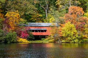 With beautiful reflections on Lake Loretta in Alley Park, Lancaster, Ohio, the red George Hutchins Covered Bridge, surrounded by colorful autumn leaves, was constructed in 1865 at another location. - 389795553