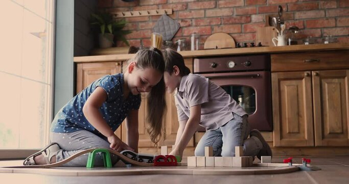 Siblings playing in home kitchen on warm floor little 7s boy build tower use wooden blocks while girl ride train on toy railroad. Hobby and leisure, fine motor skills, imagination development concept