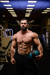 strong young bearded male with powerful abdominal muscle holding heavy training ball in dark night sport gym