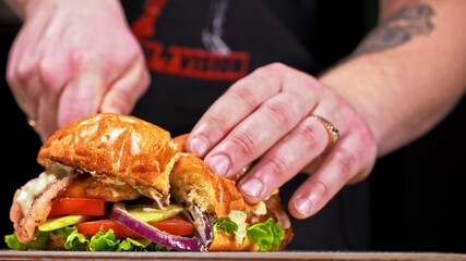 Cut craft burger is cooking on black background. Consist: sauce, lettuce, tomato, red onion, cucumber, cheese, bacon, air bun and breast of chicken. Not made ideal. Looks real, loving hand made.