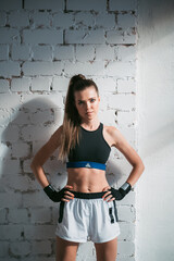 A pretty long-haired brunette of athletic build stands against a brick wall in the loft