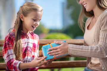 Mom giving her smiling daughter present box on park bench
