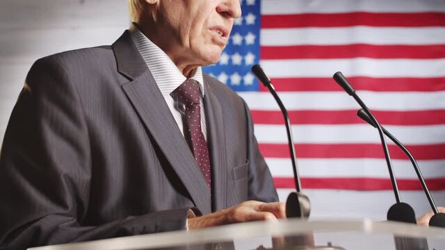 Midsection of senior male politician in formal suit standing at rostrum with microphones against U.S. flag and giving speech during press conference