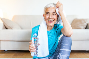 Elderly woman sitting on the mat, exhausted after the daily training. Senior woman taking a break while exercising at home. Athletic mature woman holding bottle of water