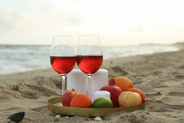Tray with glasses of wine, fruits and candles on sandy sea beach