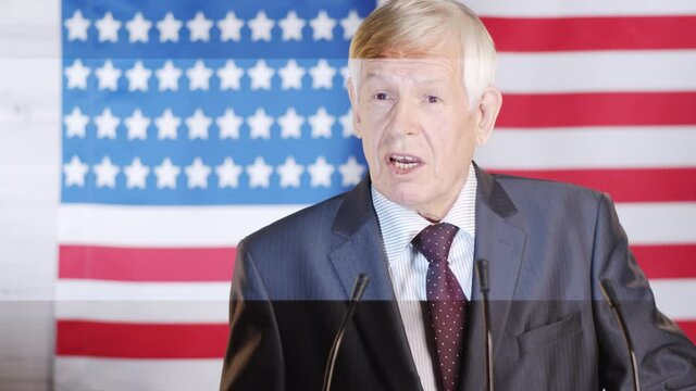 Senior Caucasian male politician in formal suit standing against U.S. flag and talking into microphones while giving public speech during press conference