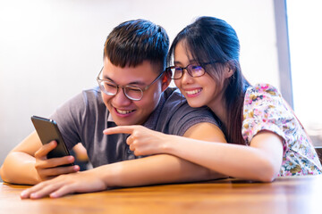 Happy asian couple in coffee shop using smartphone together, smiling young couple embracing while looking at smartphone.