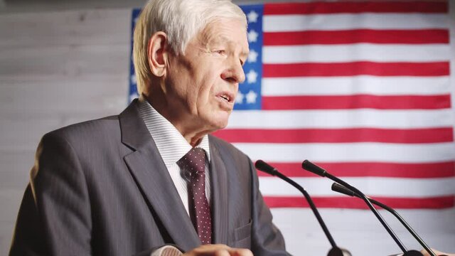 Senior male politician in formalwear giving public speech during press conference in studio with American flag in the background