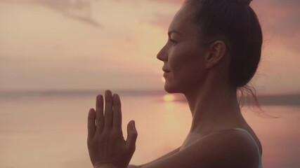 Close up of young sun-tanned Caucasian girl with hair in bun standing in wind greeting namaste looking into distance. Beautiful sky with sun setting over horizon on background