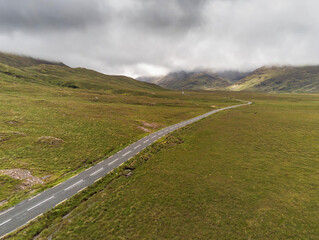 Small asphalt road into mountains. Aerial drone view, Connemara region, West of Ireland. Cloudy sky. Empty wild fields on each side of the road. Country side. Two cars on the road. Concept travel.