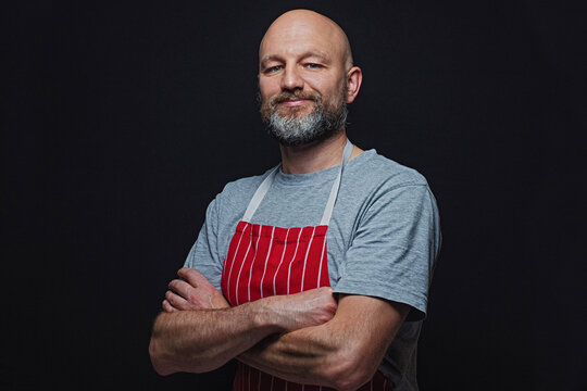 Professional fishmonger or butcher in grey t-shirt and red and white stripe classic apron on a black background. Bold male with grey beard in his 40s, Slim body type. Hands crossed Smile expression