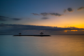 Sunrise view. Seascape. Mountains and Agung volcano. Traditional gazebos on an artificial island in the ocean. Water reflection. Sanur beach, Bali, Indonesia.