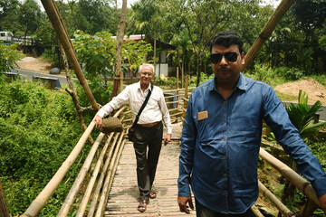 Two men are standing on a bamboo bridge in Mawlynnong, East Khasi Hills, Meghalaya, India. The young and old persons, wearing blue and white shirts, are looking towards the camera with pose and style.