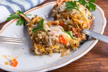 Baked eggplant with vegetables and cheese sprinkled with chopped parsley on an oval plate on a wooden table with a fork and knife, close-up