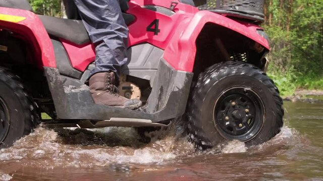Tracking shot of unrecognizable man driving red quad bike through shallow river in forest