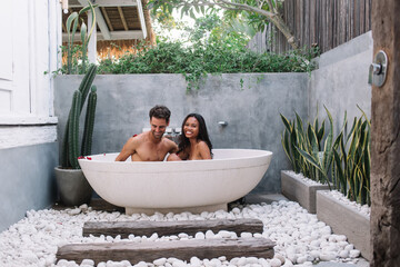 Smiling multiracial couple in love recreating on resort during honeymoon sitting in bathtub outdoors with rose petals, cheerful male and female relaxing on spa aromatherapy together on vacation
